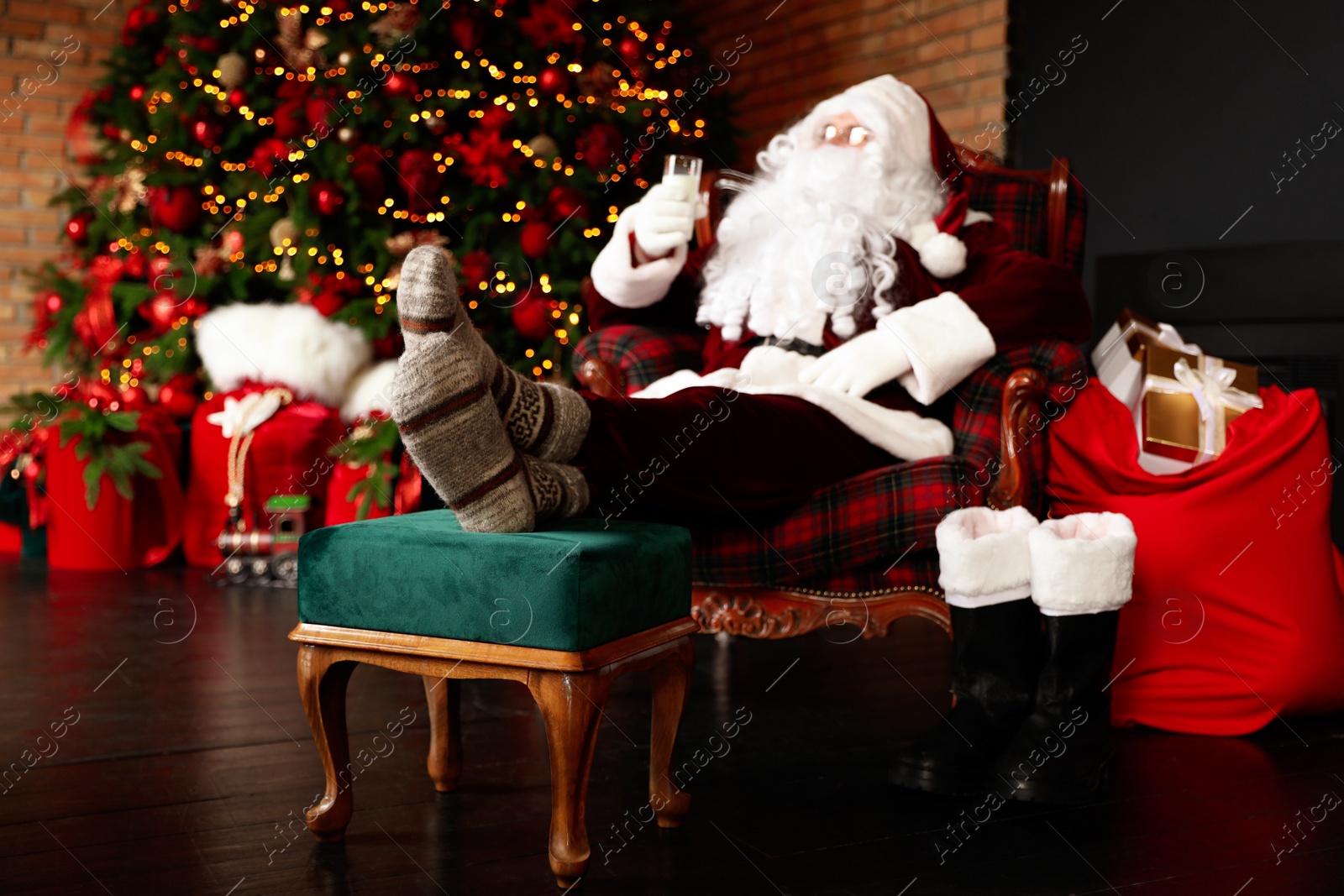 Photo of Santa Claus with glass of milk resting in armchair near Christmas tree, focus on legs
