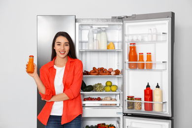 Happy young woman with juice near open refrigerator indoors