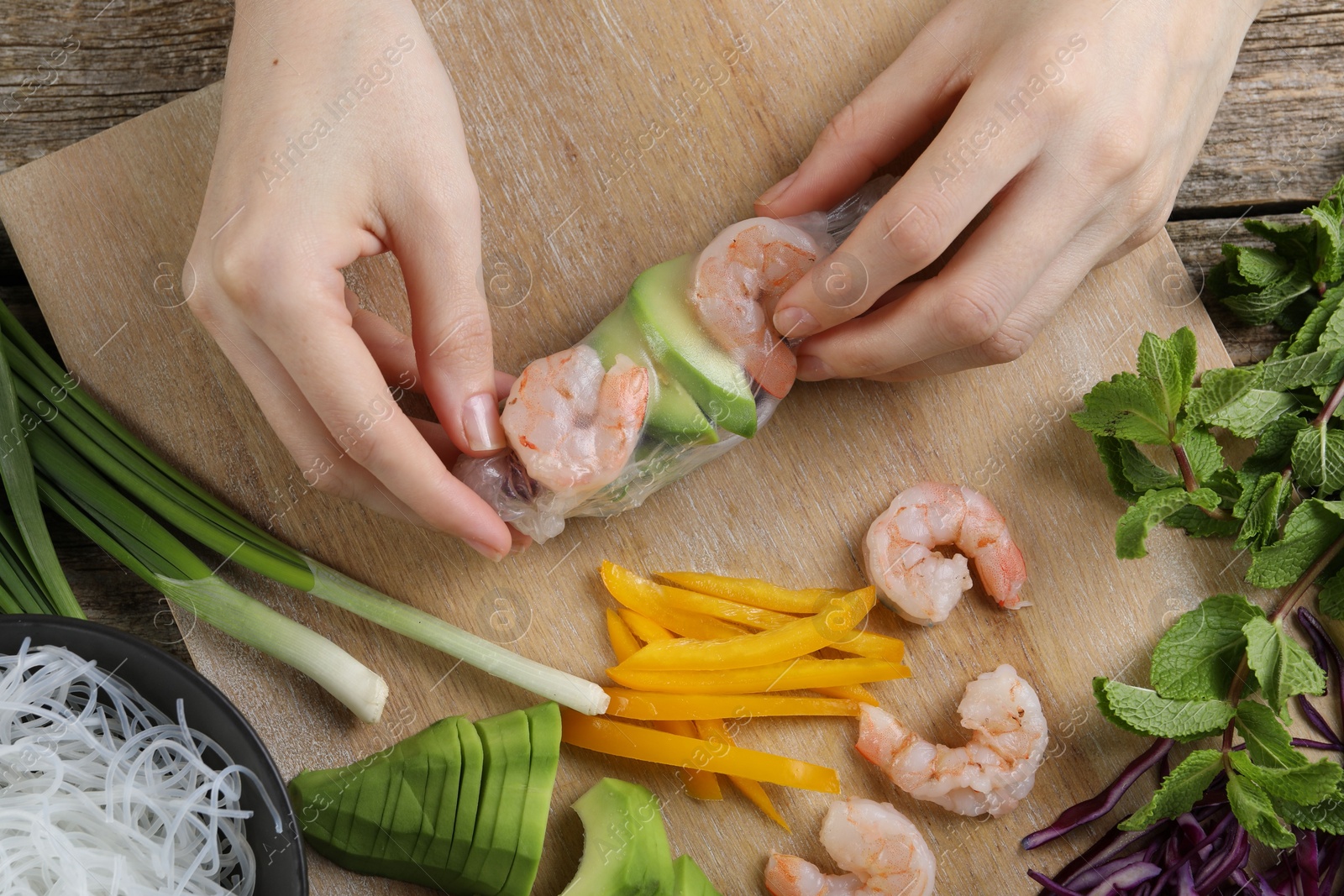 Photo of Woman wrapping spring roll at wooden table with products, top view