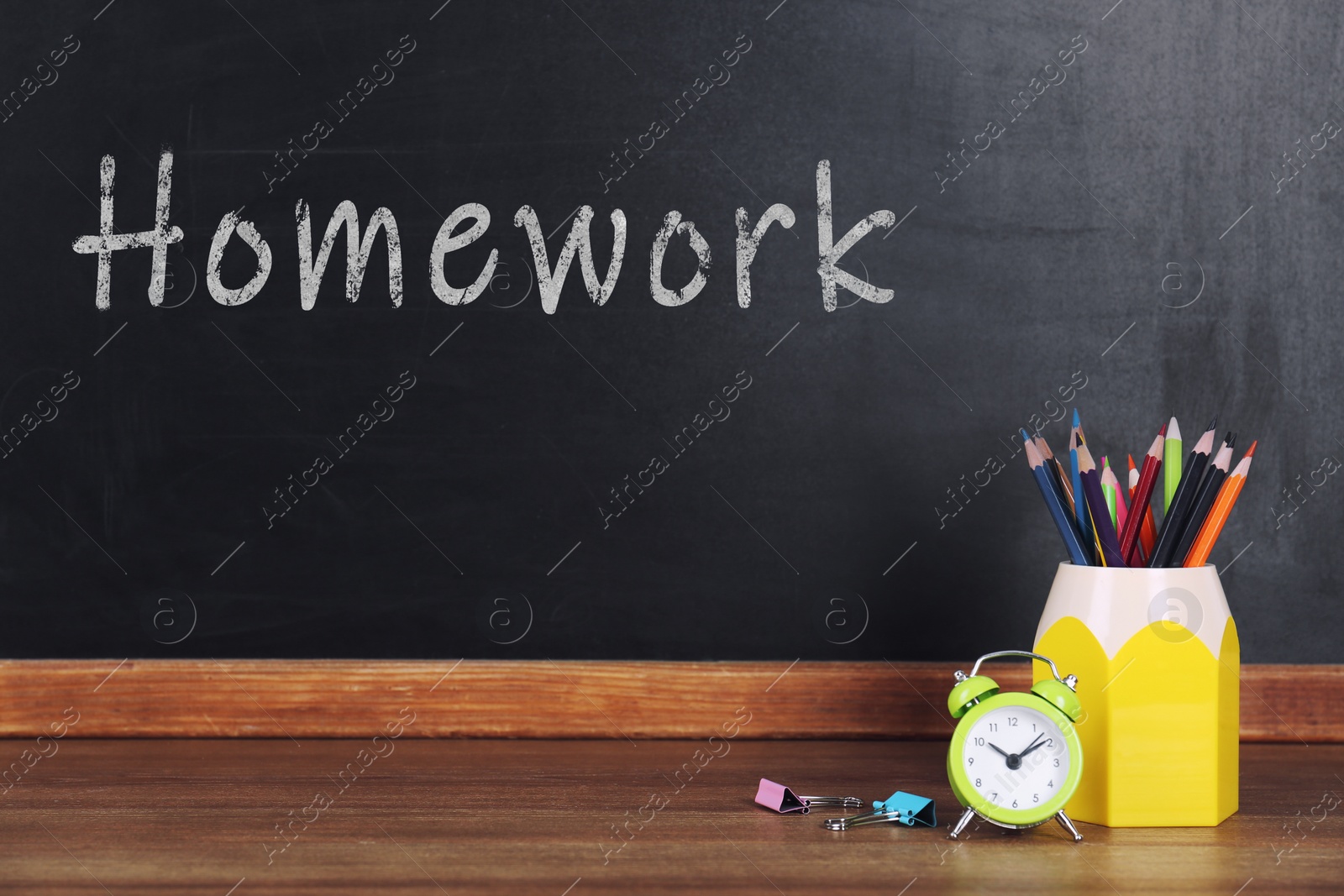 Image of Pencils and alarm clock on wooden table near blackboard. Doing homework