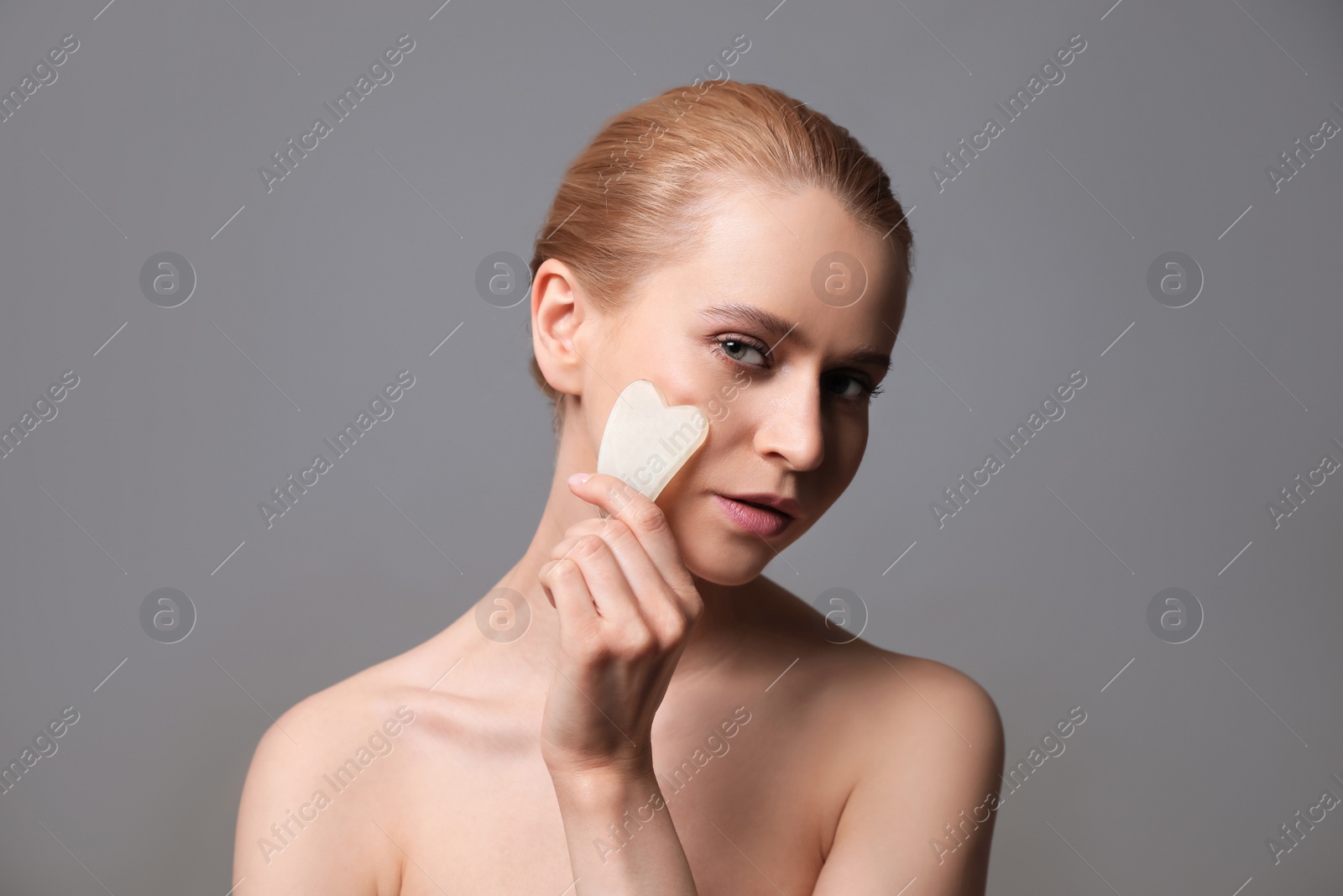 Photo of Beautiful young woman doing facial massage with gua sha tool on grey background