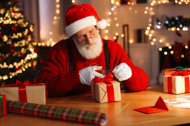 Photo of Santa Claus tying bow on gift box at his workplace in room decorated for Christmas