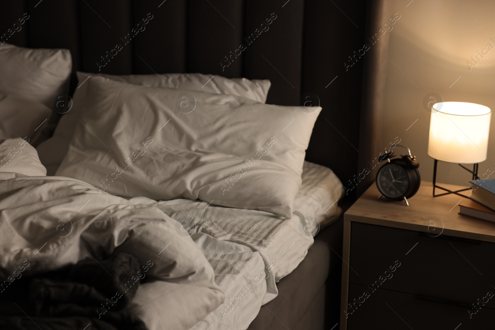 Photo of Nightlight, alarm clock, glasses and books on bedside table near bed indoors