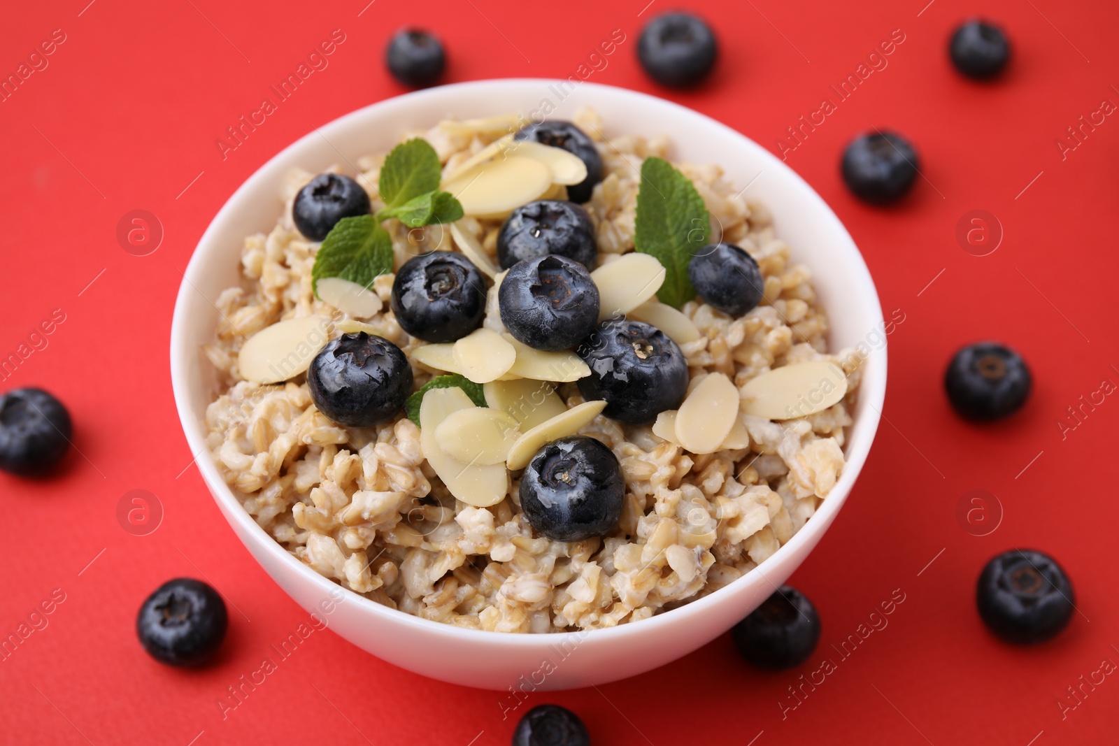 Photo of Tasty oatmeal with blueberries, mint and almond petals in bowl surrounded by fresh berries on red background