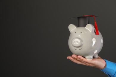 Man holding piggy bank and graduation cap against black background, closeup with space for text. Scholarship concept