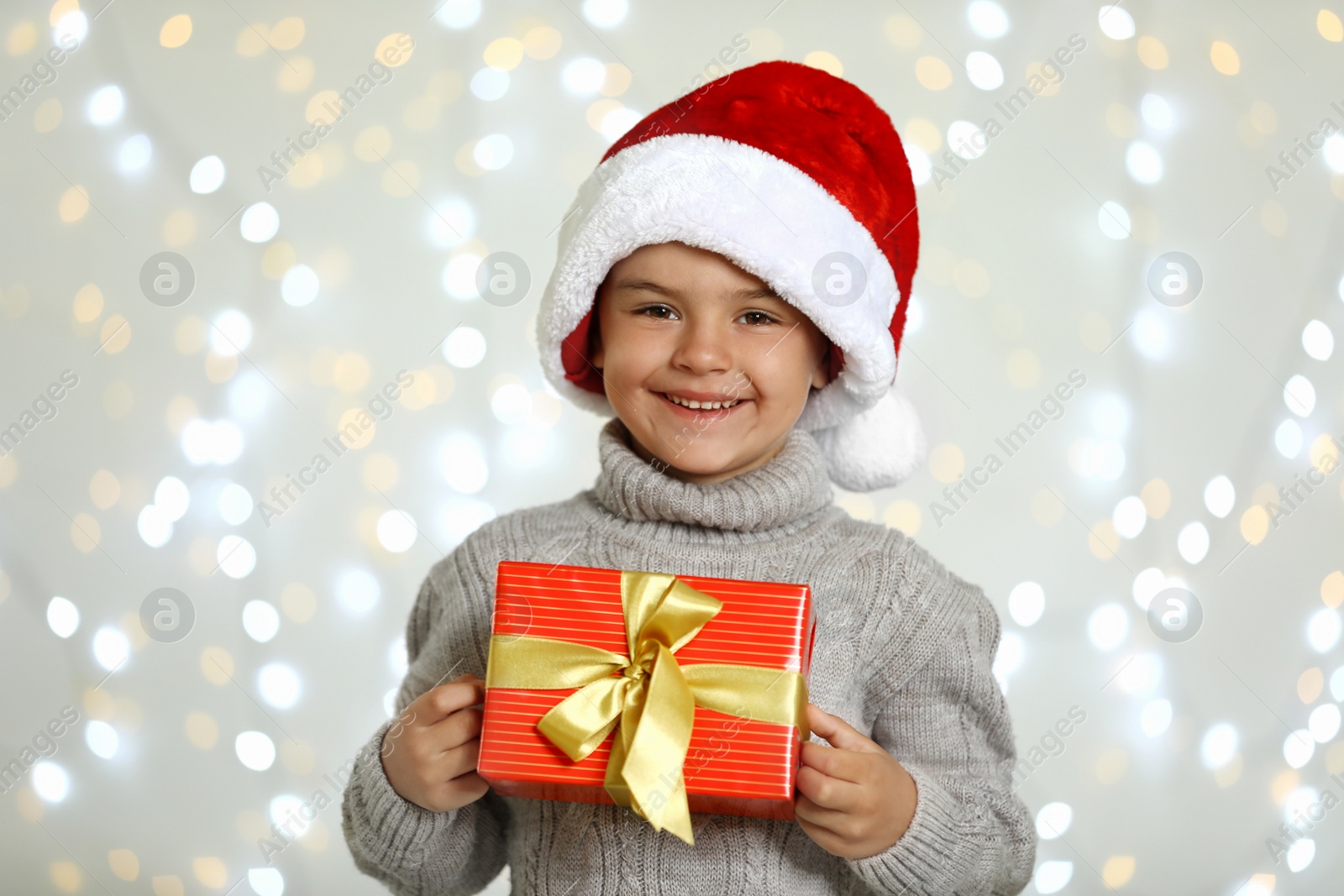 Photo of Happy little child in Santa hat with gift box against blurred festive lights. Christmas celebration