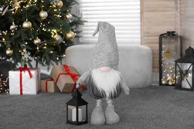 Photo of Cute Scandinavian gnome with lantern candle holder near Christmas tree on carpet in room