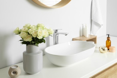 Vase with beautiful white roses and toiletries near sink in bathroom