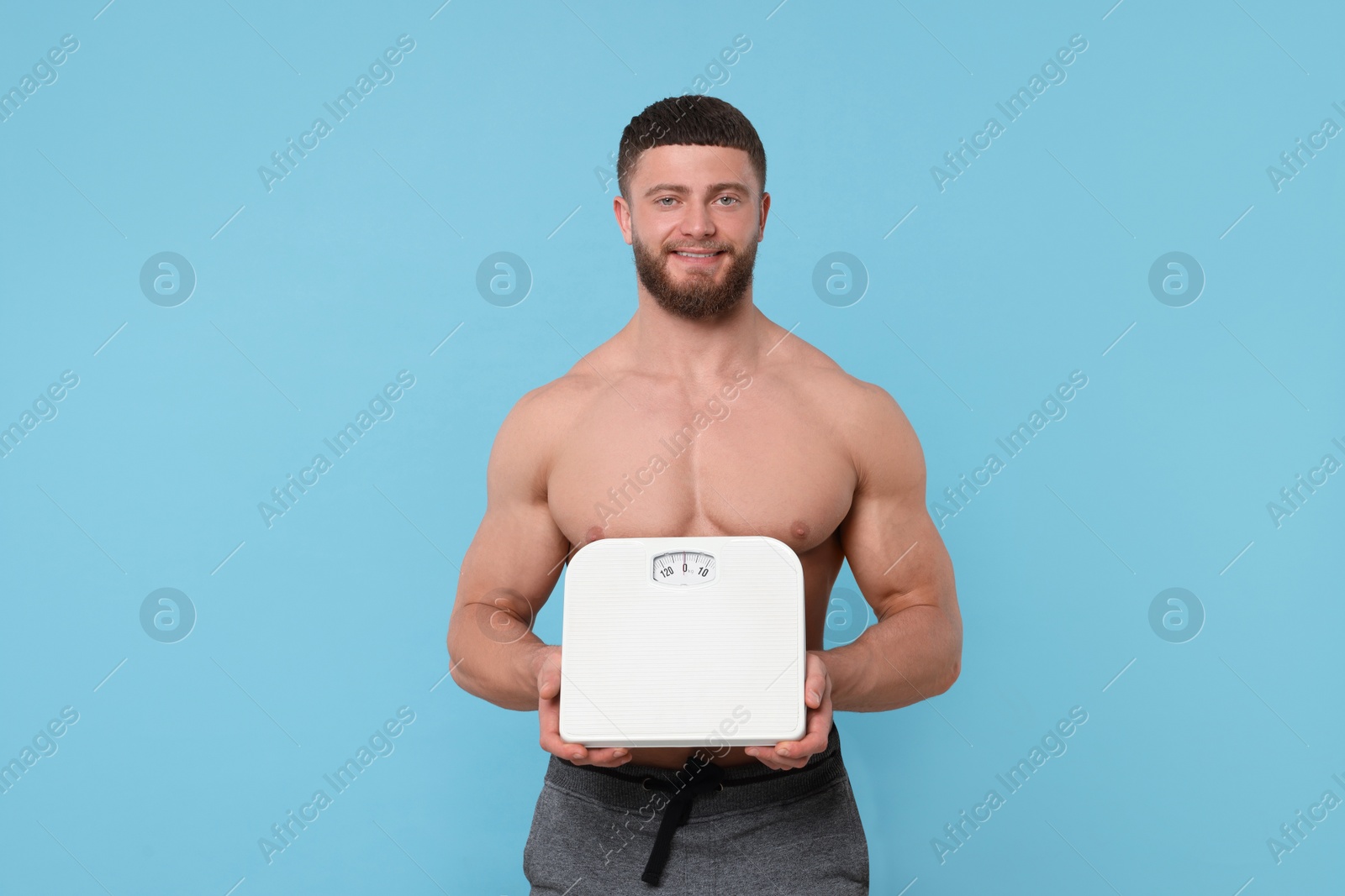 Photo of Happy athletic man holding scales on light blue background. Weight loss concept