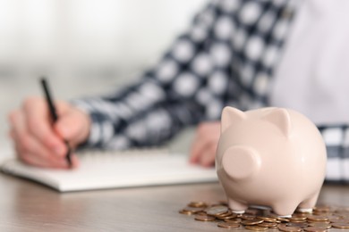 Photo of Financial savings. Man writing down notes at wooden table, focus on piggy bank and coins