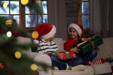 Photo of Happy children wearing Santa hats with Christmas gifts on sofa at home