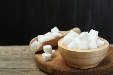 Photo of White sugar cubes in bowl and scoop on wooden table against dark background, closeup. Space for text