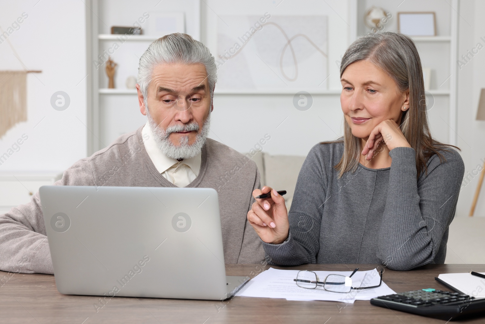 Photo of Elderly couple with papers and laptop discussing pension plan at wooden table in room