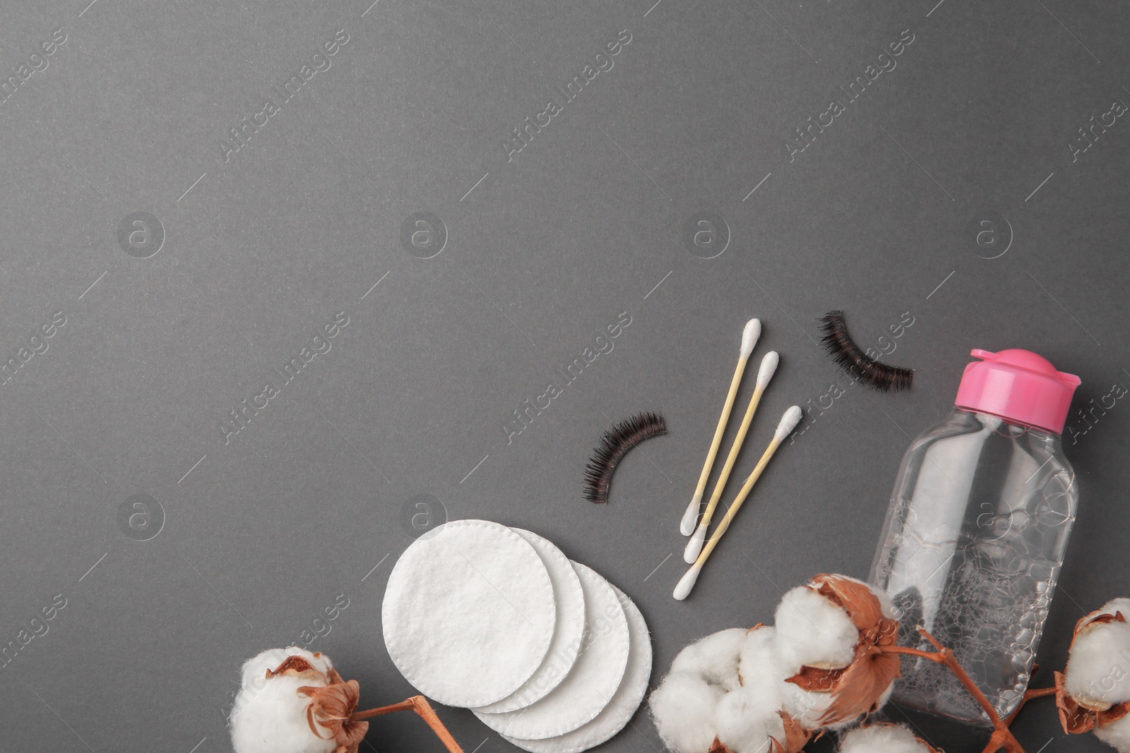 Photo of Bottle of makeup remover, cotton flowers, pads, swabs and false eyelashes on grey background, flat lay. Space for text