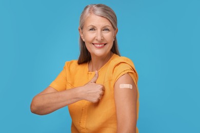 Photo of Senior woman with adhesive bandage on her arm after vaccination showing thumb up against light blue background