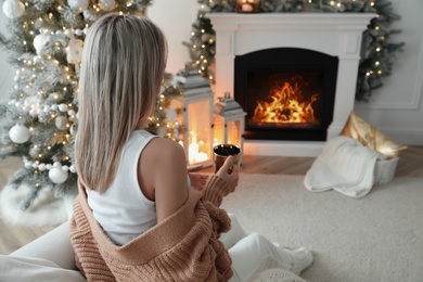 Woman with hot drink resting near fireplace in cozy room decorated for Christmas, back view