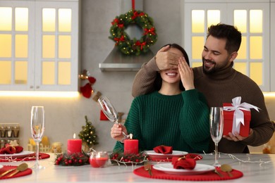 Happy young man surprising his girlfriend with Christmas gift at table in kitchen