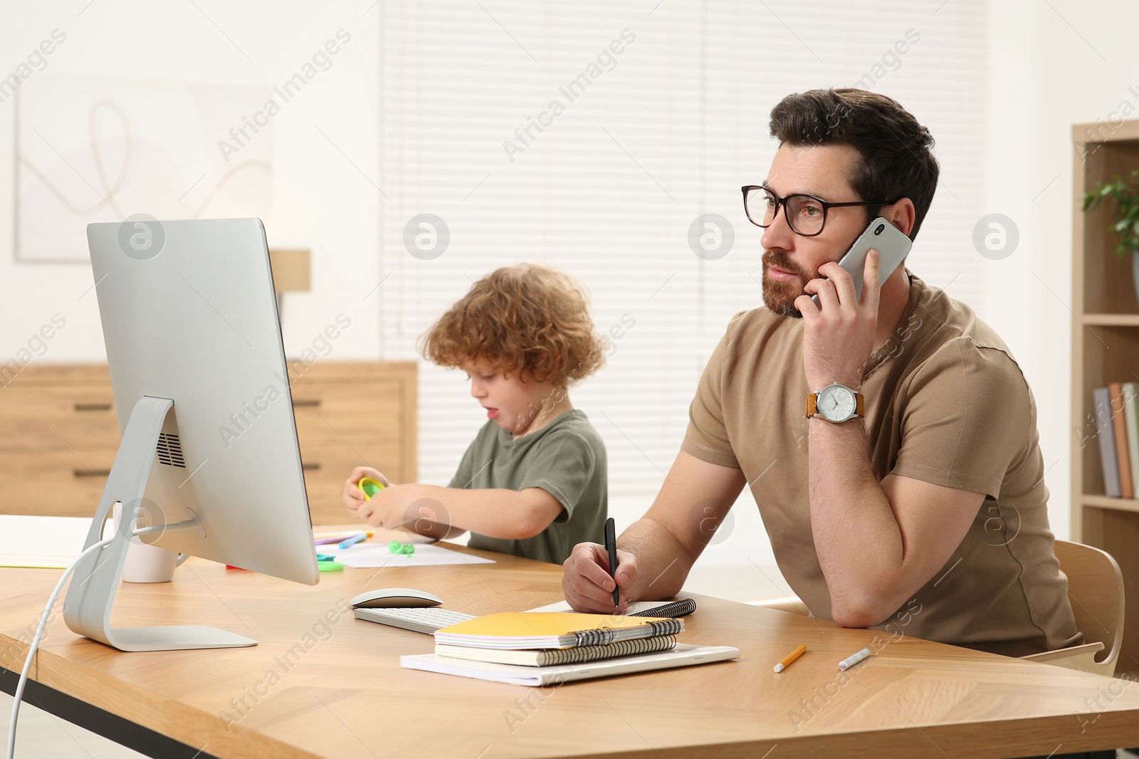 Photo of Man working remotely at home. Busy father talking on smartphone while his son using play dough at desk