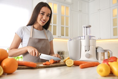 Young woman cutting fresh carrot for juice at table in kitchen