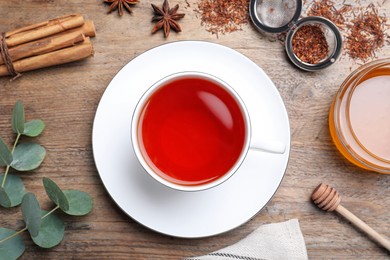 Photo of Freshly brewed rooibos tea, scattered dry leaves, honey and spices on wooden table, flat lay