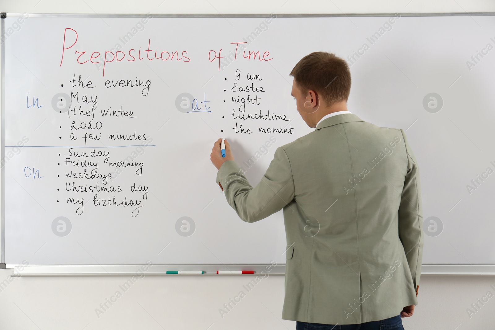 Photo of English teacher giving lesson on prepositions of time near whiteboard in classroom, back view