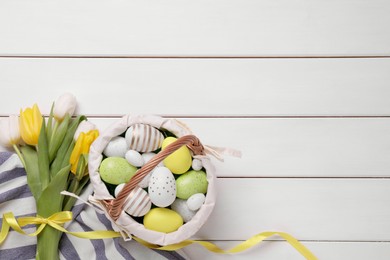 Photo of Easter eggs in basket and tulips on white wooden table, flat lay. Space for text