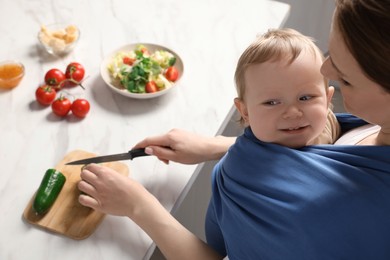 Mother cutting cucumber while holding her child in sling (baby carrier) indoors, above view