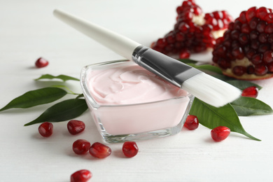 Photo of Glass bowl with natural facial mask, pomegranate seeds and brush on white wooden table, closeup