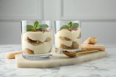 Photo of Delicious tiramisu in glasses, mint leaves, cookies and spoons on white marble table