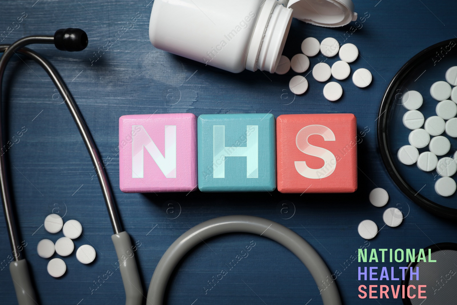 Image of National health service. Abbreviation NHS made with cubes, stethoscope and pills on blue wooden background, flat lay