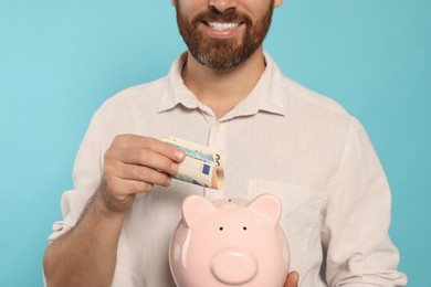 Happy man putting money into piggy bank on light blue background, closeup