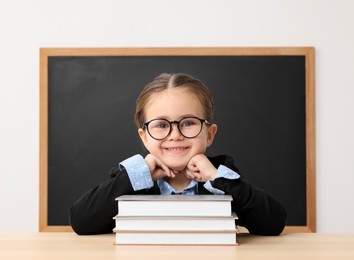 Happy little school child sitting at desk with books near chalkboard in classroom