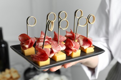 Photo of Woman holding plate of tasty canapes with ham and cheese indoors, closeup