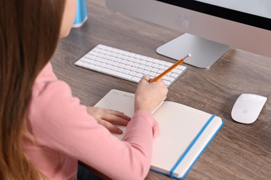 Photo of E-learning. Girl taking notes during online lesson at table indoors, closeup