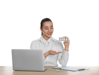 Professional pharmacist with pills and laptop at table against white background