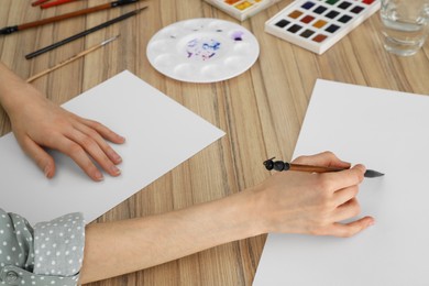 Photo of Woman painting with watercolor on blank paper at wooden table, closeup
