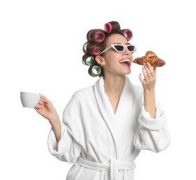 Beautiful young woman in bathrobe with hair curlers holding croissant and cup of drink on white background