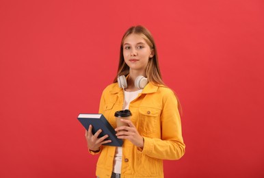 Teenage student with book, cup of coffee and headphones on red background