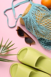 Photo of String bag with towel, sunglasses, orange, slippers, cream and palm leaf on pink background, flat lay