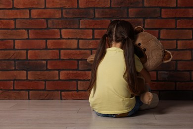 Photo of Child abuse. Upset little girl with teddy bear sitting on floor near brick wall indoors, back view. Space for text