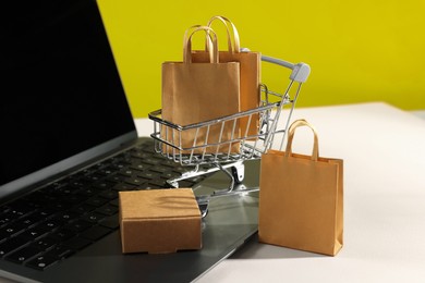Photo of Online store. Laptop, mini shopping cart and purchases on beige table, closeup