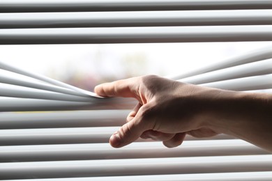 Photo of Man separating slats of white blinds indoors, closeup