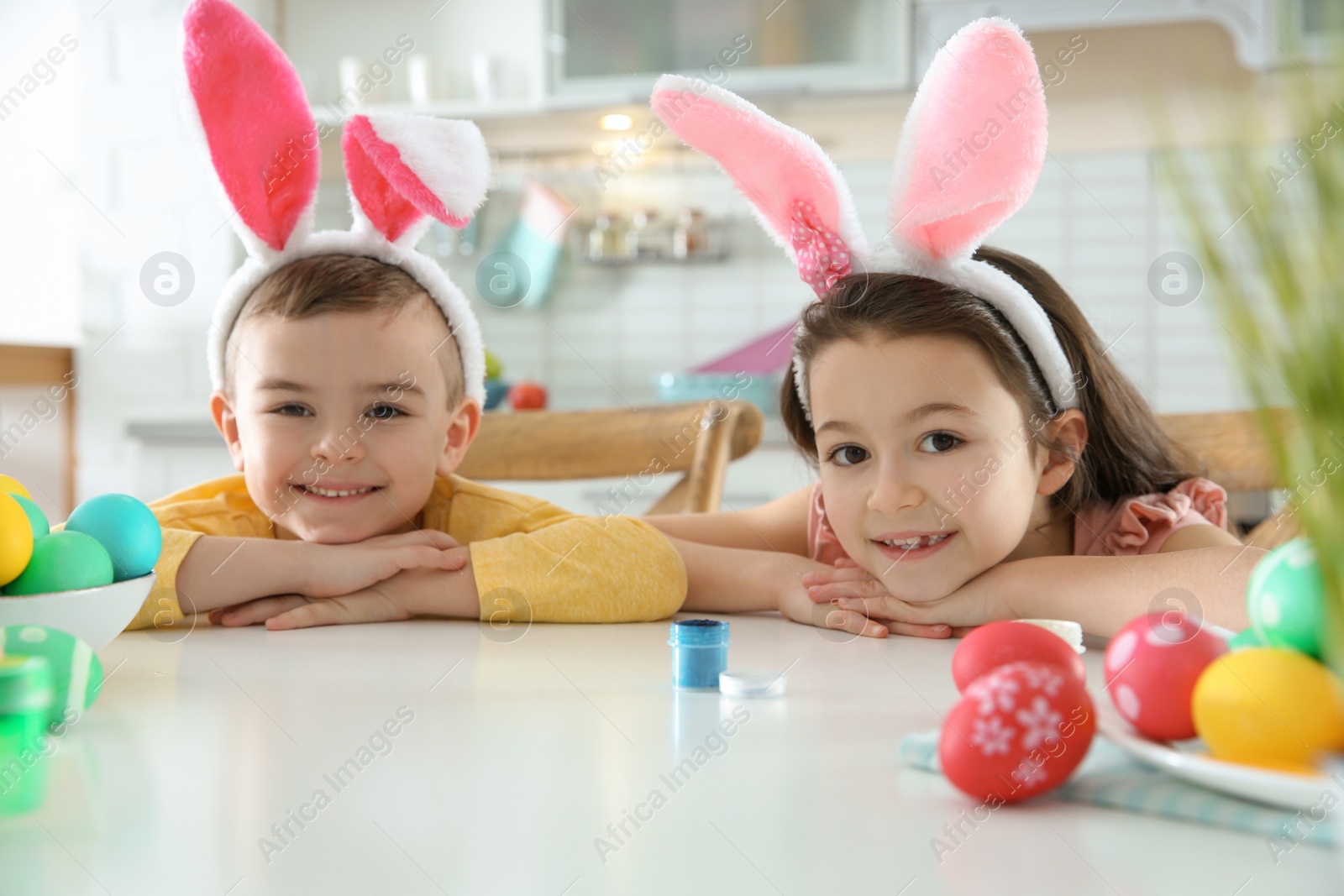 Photo of Cute children with bunny ears headbands and painted Easter eggs sitting at table in kitchen