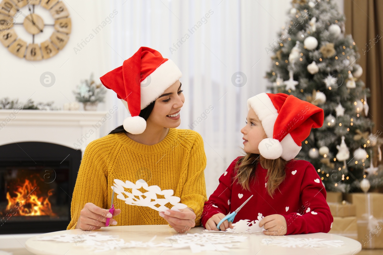 Photo of Mother and daughter in Santa hats making paper snowflakes near Christmas tree at home