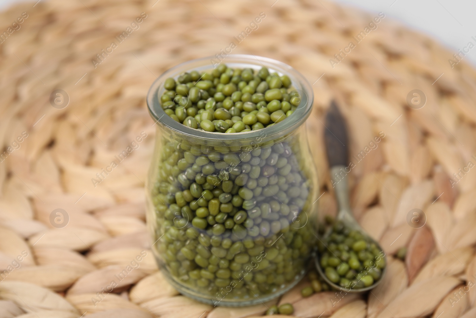 Photo of Glass jar of mung beans with spoon on wicker mat, closeup