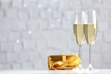 Sparkling wine in glasses near gift box on table against blurred lights, space for text
