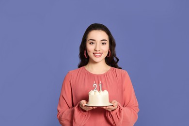 Coming of age party - 21st birthday. Smiling woman holding delicious cake with number shaped candles on violet background