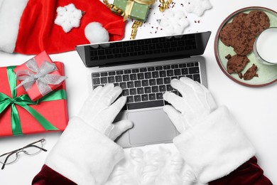 Santa Claus using laptop, closeup. Gift boxes, eyeglasses and biscuits on white table, top view