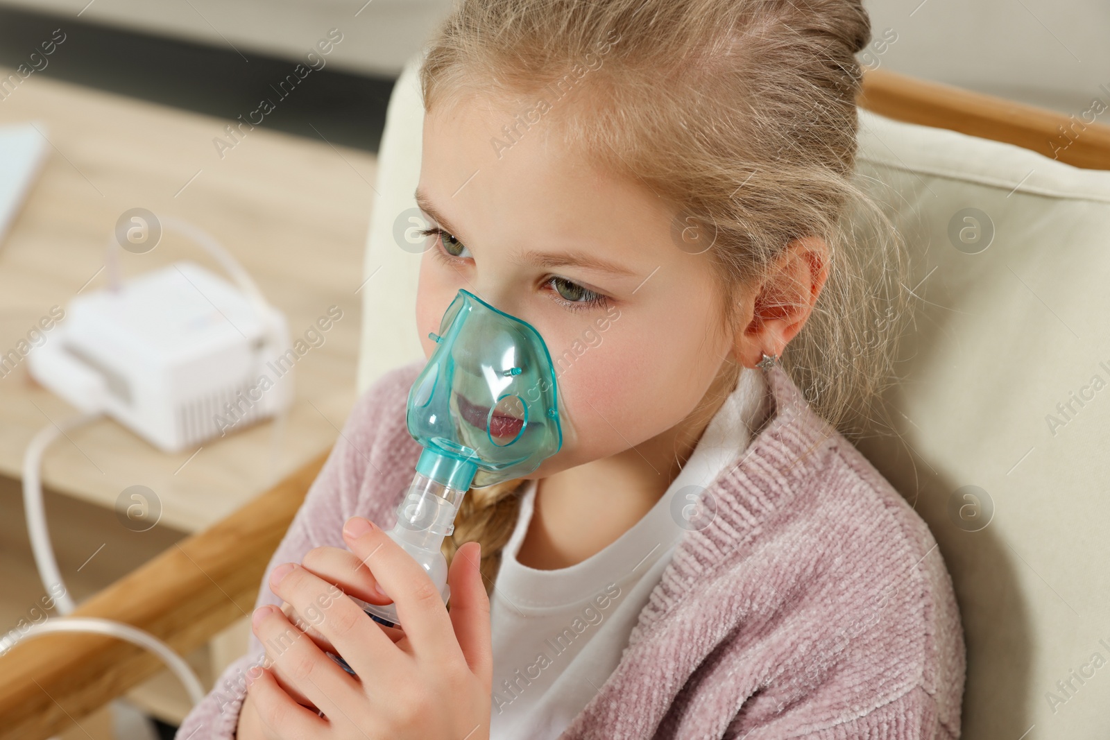 Photo of Little girl using nebulizer for inhalation in armchair at home