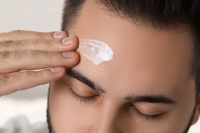 Man with dry skin applying cream onto his forehead on light background, closeup